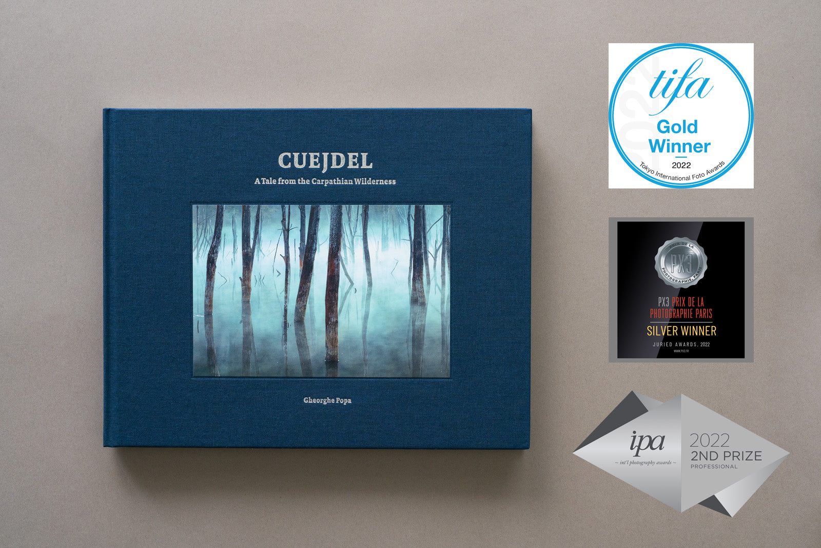 Vedere de sus a albumului foto „Cuejdel - A Tale from the Carpathian Wilderness” de Gheorghe Popa, cu copertă textilă albastră și o fotografie centrală cu arbori în ceață. Lângă album sunt afișate trei embleme de premii internaționale: TIFA Gold Winner 2022, PX3 Silver Winner 2022 și IPA 2nd Prize Professional 2022.