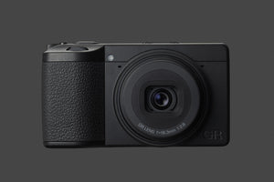 Detaliu frontal Ricoh GR IV Monochrom pe fundal gri, evidențiind grip-ul texturat, obiectivul fix f/2.8 și designul minimalist de tip "stealth".