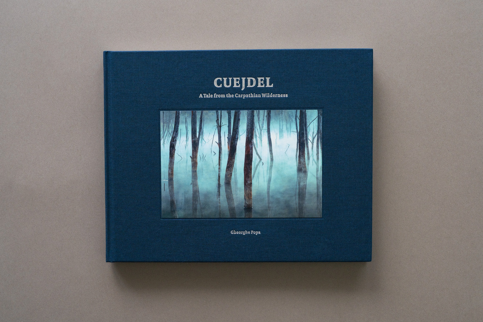 Vedere frontală a albumului de artă fotografică Cuejdel, realizat de Gheorghe Popa. Cartea are o copertă din material textil de culoare albastru închis, cu titlul gravat și fotografia Cold and Misty aplicată central pe copertă ce înfățișează trunchiuri de copaci scufundați în lacul Cuejdel, înconjurați de o ceață densă și misterioasă.