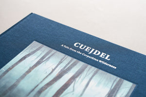 Prim-plan detaliat (macro) al copertei albumului foto „Cuejdel - A Tale from the Carpathian Wilderness”. Imaginea evidențiază textura fină a materialului textil albastru și literele albe, reliefate, ale titlului, sugerând calitatea premium a tiparului și a legătoriei de artă.