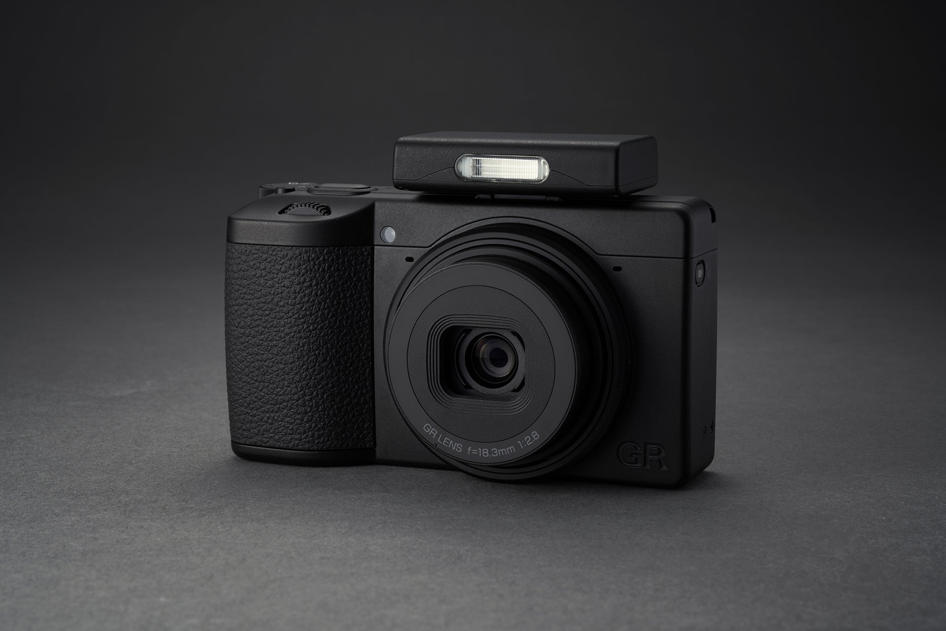 Aparat foto Ricoh GR IV Monochrom cu bliț extern compact montat pe patina superioară, prezentat pe fundal gri antracit pentru evidențierea texturilor.
