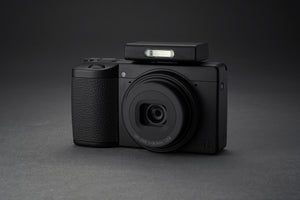 Aparat foto Ricoh GR IV Monochrom cu bliț extern compact montat pe patina superioară, prezentat pe fundal gri antracit pentru evidențierea texturilor.