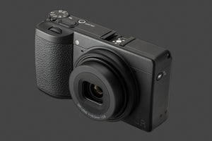 Vedere de sus a aparatului Ricoh GR IV Monochrom, prezentând rotița de selecție a modurilor, butonul de pornire și patina pentru accesorii pe un corp compact negru.