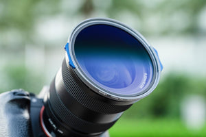 Cadru de detaliu cu filtrul gradual magnetic NiSi JetMag Pro Medium FS GND8 montat pe un obiectiv grandangular, evidențiind marginile albastre de prindere și tratamentul sticlei, pe un fundal natural cu bokeh verde.