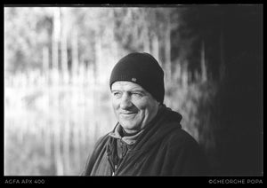 Portret alb-negru pe film Agfa APX 400 cu un bărbat zâmbitor purtând căciulă și jachetă cu glugă.