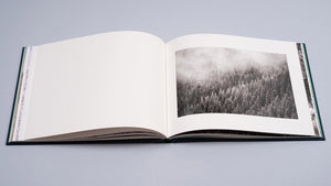 Albumul foto „Cuejdel” de Gheorghe Popa, deschis larg pentru a evidenția designul editorial curat. Pagina din dreapta conține o fotografie monocromă cu o pădure de brazi acoperită de nori și ceață, în timp ce pagina din stânga rămâne albă, creând un echilibru vizual ce pune în valoare detaliile fine ale peisajului carpatin.